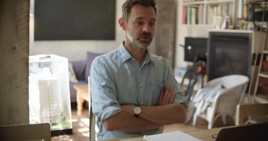 A middle aged man writes on a sheet of paper while using a laptop to participate in a video conference