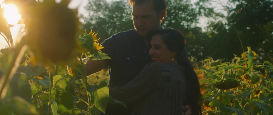 A man and woman embrace while examining a sunflower in a field of sunflowers during the day