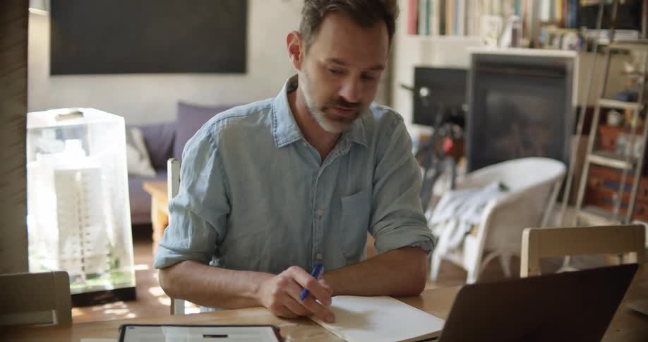 A man writes in a notebook and taps the screen of a tablet while participating in a video chat on a laptop as he sits at a desk in the living room of a house during the day