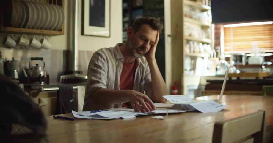 A man goes over paper bills while sitting at his kitchen table