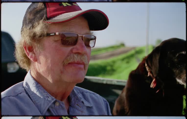 Man wearing baseball cap and sunglasses sitting in a car with his dog in a rural area