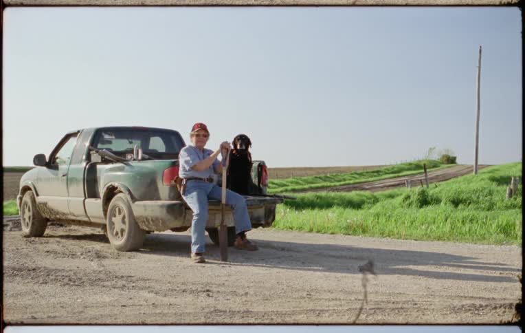 Man holding a shovel sitting with his dog on the back of a truck parked in a rural road
