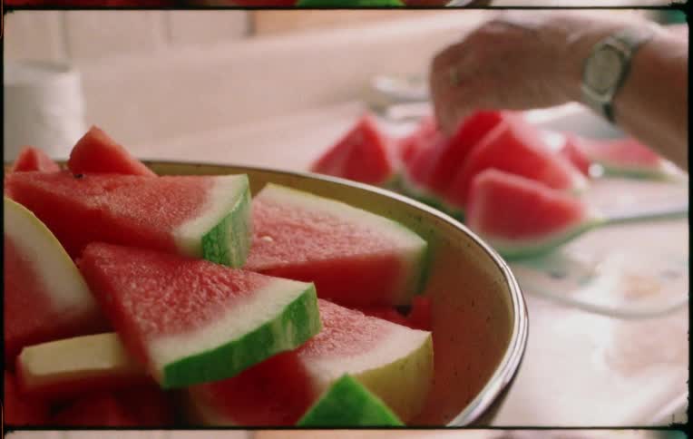 A person in a home kitchen cuts up watermelon into wedges and places it in a bowl on a countertop