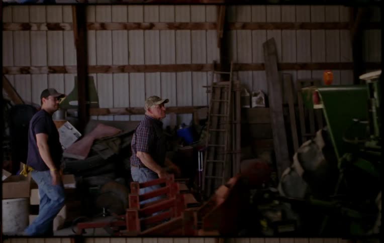 A farmer opens the door of a warehouse and climbs up over a tractor parked inside