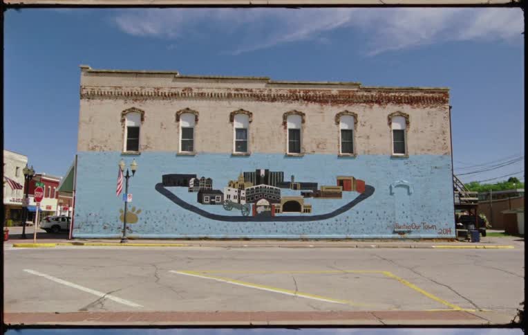 A city street with a car driving by a building with buildings the sky and clouds in the background