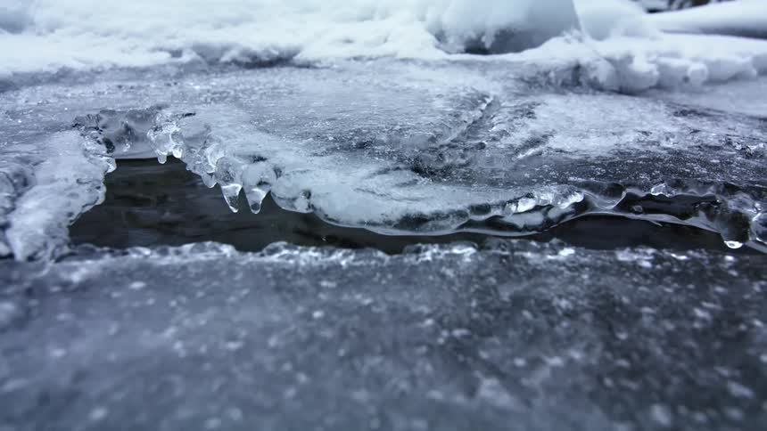 A stream of water flows beneath a sheet of ice outdoors during the day