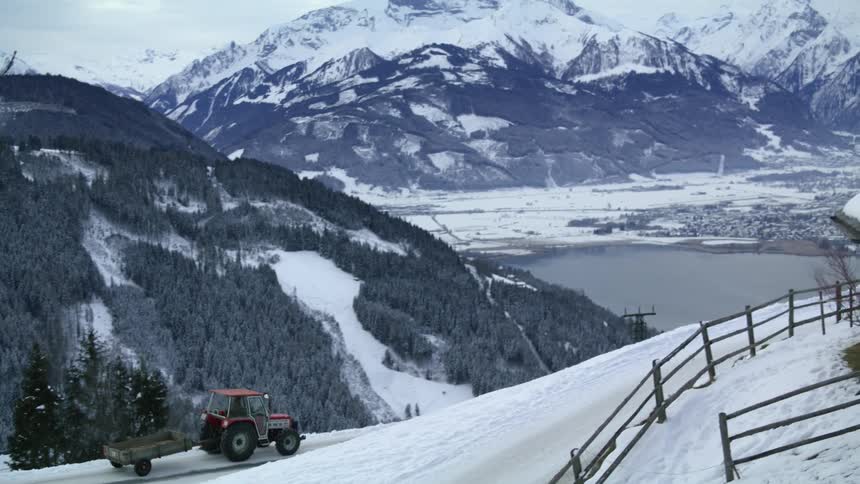 A vehicle with a trailer driving on snow with a mountain range the sky and clouds in the background