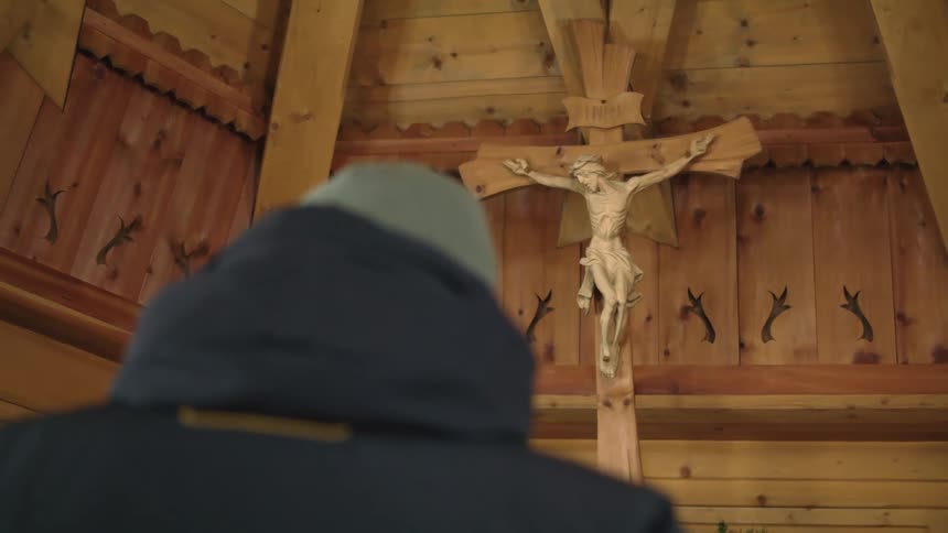 Person wearing winter coat bows head in front of cross in wooden walled church