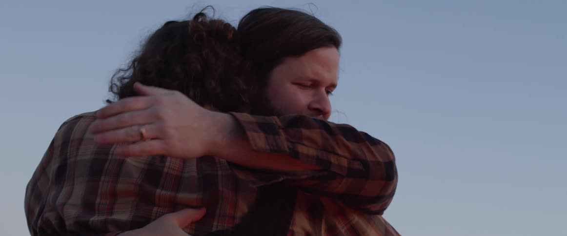 Two friends wearing flannel shirts greet each other with a hug outdoors at sunset