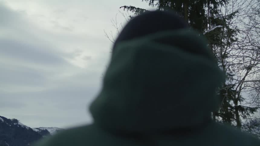 A man with a hoodie and a hat standing with a mountain range the sky and clouds in the background