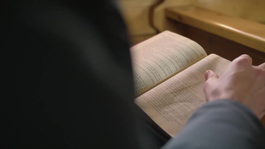 Hands open a bible and turn the pages as a person sits in a pew in a church
