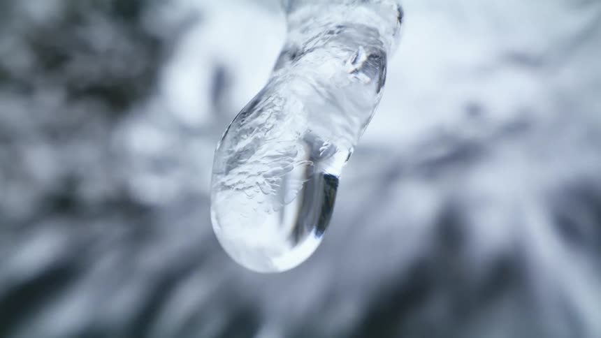 Water drop dripping from an icicle hanging near a waterfall at winter