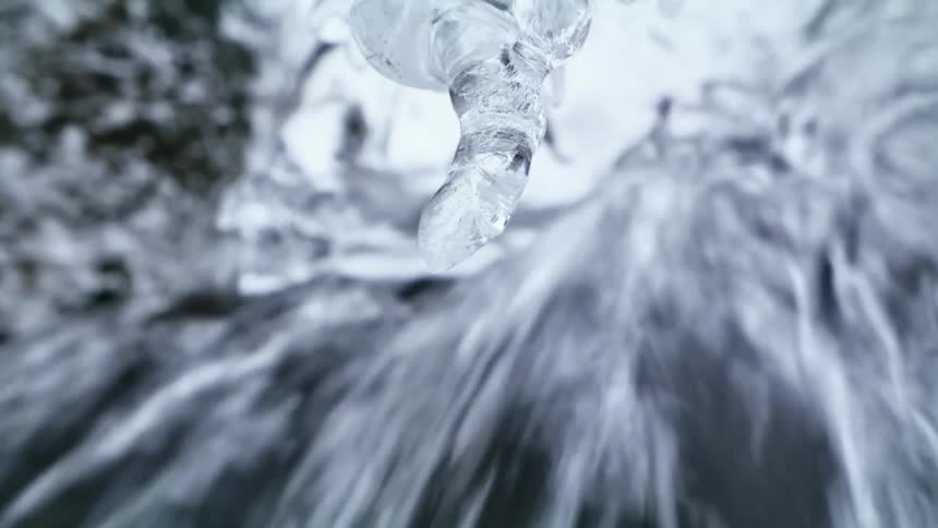 Water drop dripping from an icicle hanging near a waterfall at winter