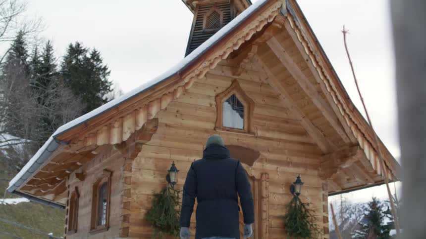 A person with a hat and coat and gloves walking towards a church with trees the sky and clouds in the background