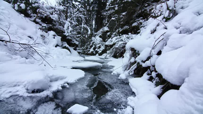 A small stream of water flows through a frozen river surrounded by sloped blankets of snow
