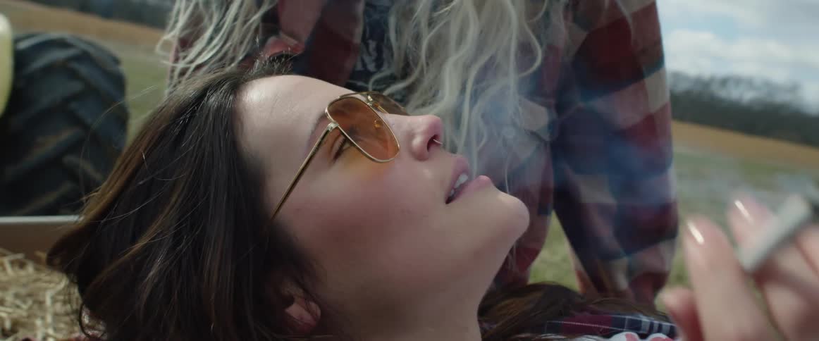 A woman smokes a cigarette and she lies down in a tractor trailer filled with hay