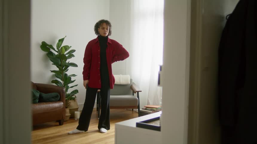 A woman standing in a living room sends a text on her phone then looks around with an upset expression on her face