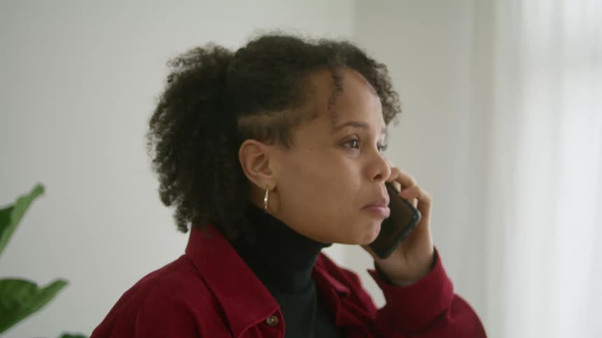 A woman in a room talking on a smart phone with a window and a wall in the background
