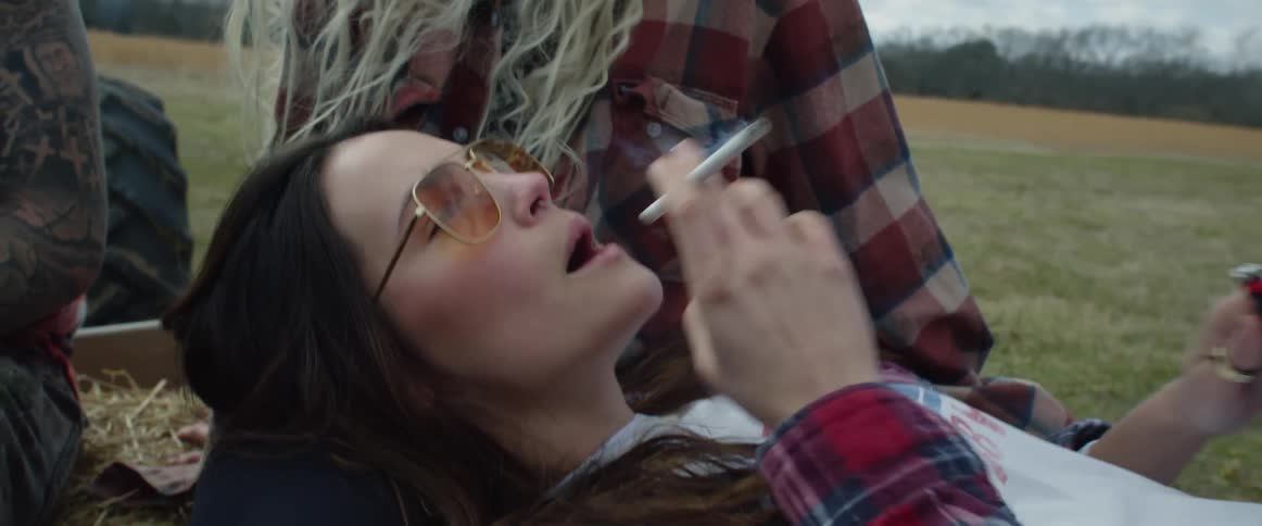 A teenage girl lies in her friends lap on a wagon and laughs as she smokes a cigarette and blows smoke at her friend