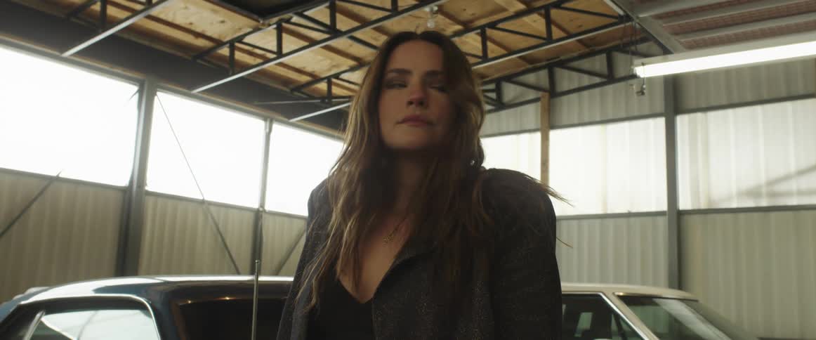 Young long haired woman modeling among the parked cars inside a warehouse looking at the camera