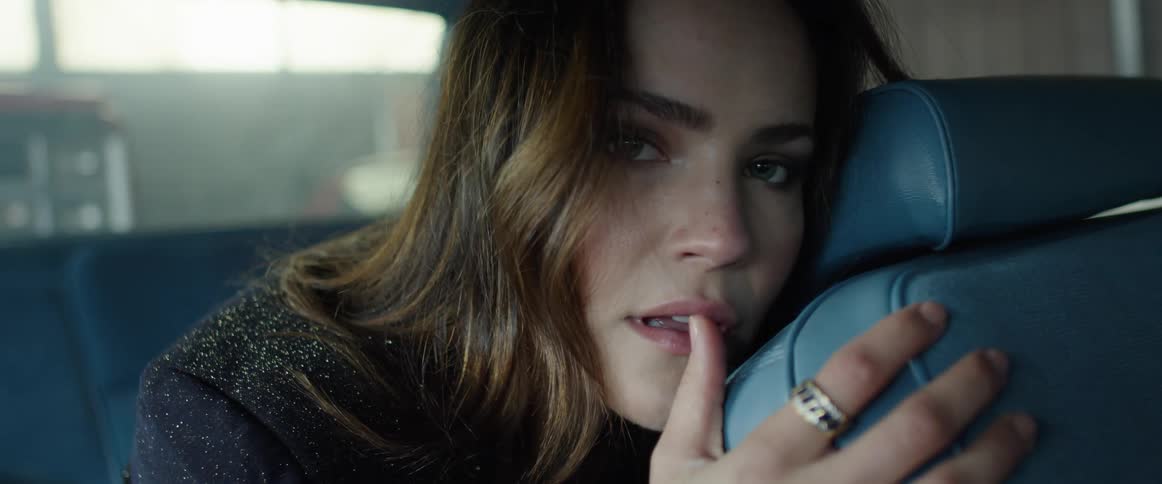 A woman sitting in the back seat of a car hugs the front seat and touches her mouth provocatively