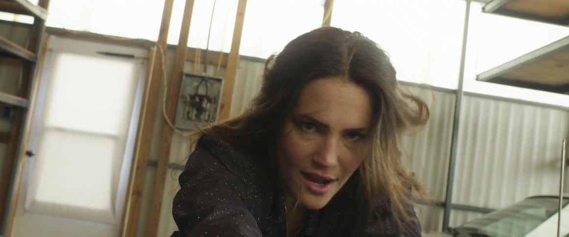 A woman in a sparkling jacket looks into the camera while leaning over the hood of a car in a garage during the day