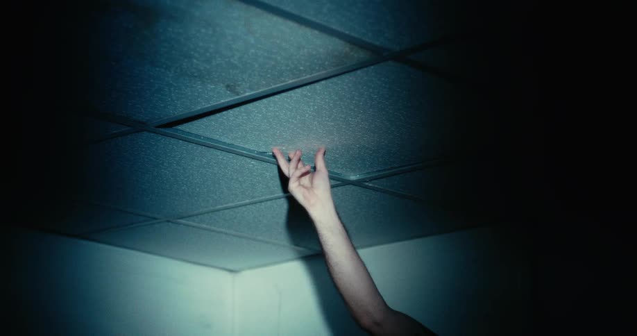 A hand lifting up a tile on the ceiling of a dimly lit room