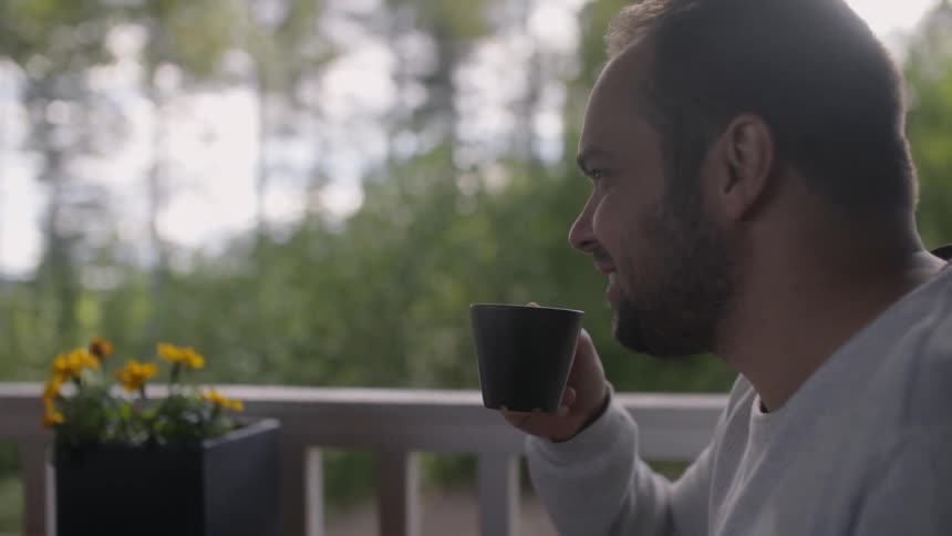 A man speaking with someone and sipping from a mug while sitting on a balcony