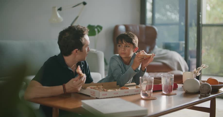 A father and son are sitting in a living room eating pizza