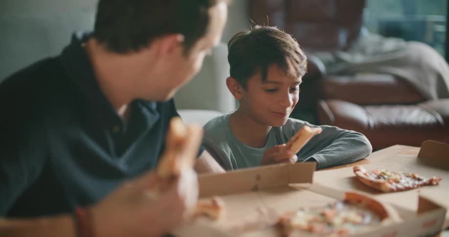 A father and son sitting at a table in a living room eating delivery pizza from a box