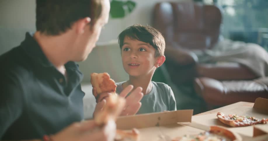 A boy gestures with the slice of pizza he is holding then takes a bite