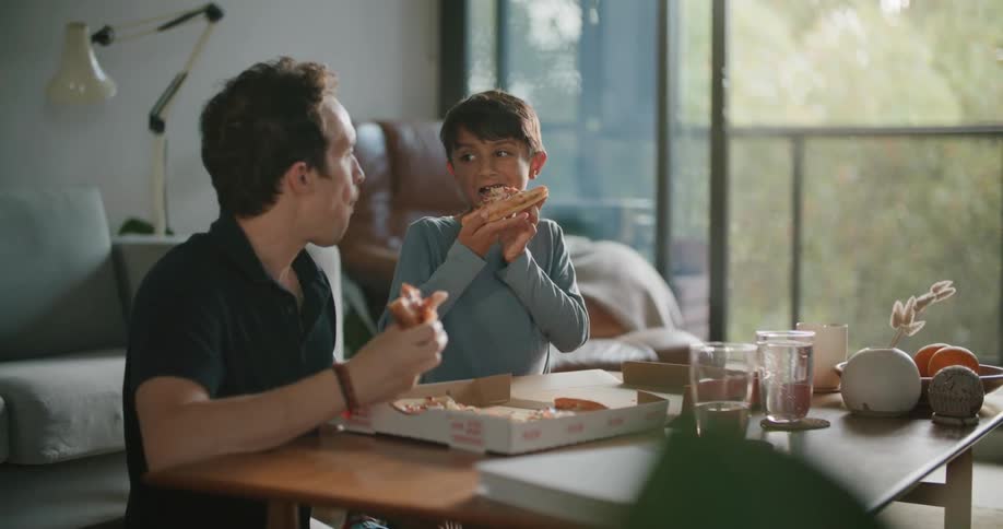 A father and son eat pizza out of a box at home
