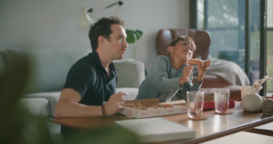 A father and son are eating pizza in their living room