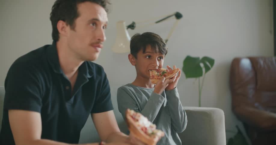 A man and a boy sit on a sofa eating pizza from a delivery box which is on the coffee table