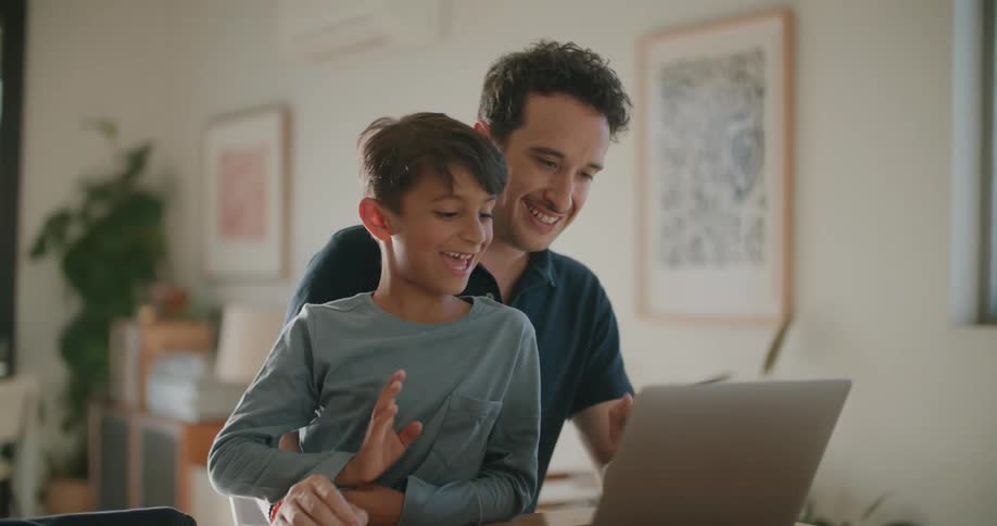 A father and son at home wave and chat with someone on a laptop