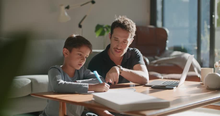A man helps his son with homework as they sit at the coffee table in their living room
