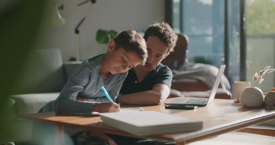 A father sits at home with his son as he does his homework