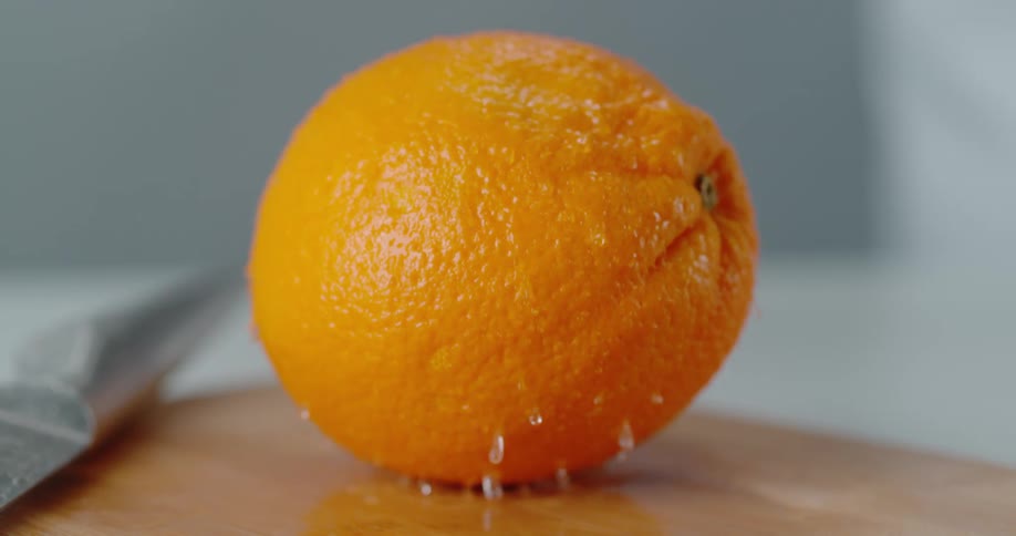 An orange lands on a cutting board and water droplets fall off of it as it bounces