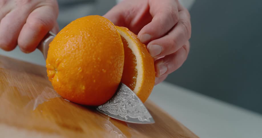 A person is holding an orange steady with one hand and using a knife to slice it in half with the other hand