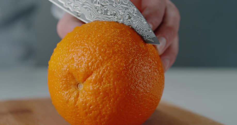 A person is holding an orange steady with their fingers as a knife blade slices it in half