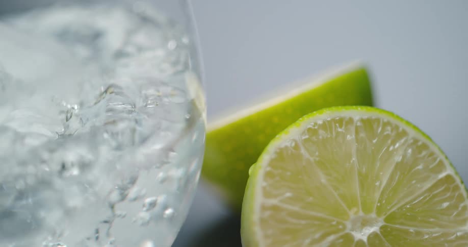 An orange and lemon next to a glass with ice with clear liquid being poured into it