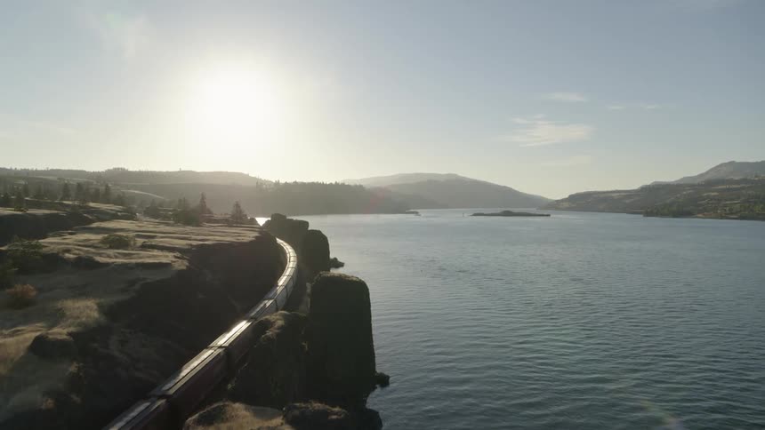 A freight train travels quickly through a narrow path of carved rock along the shore of a wide river under the morning sun