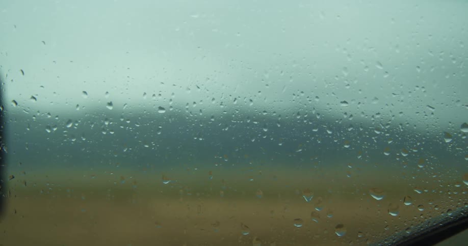 Rain drops gather and shake on a car window as it travels past grassy fields with hills behind