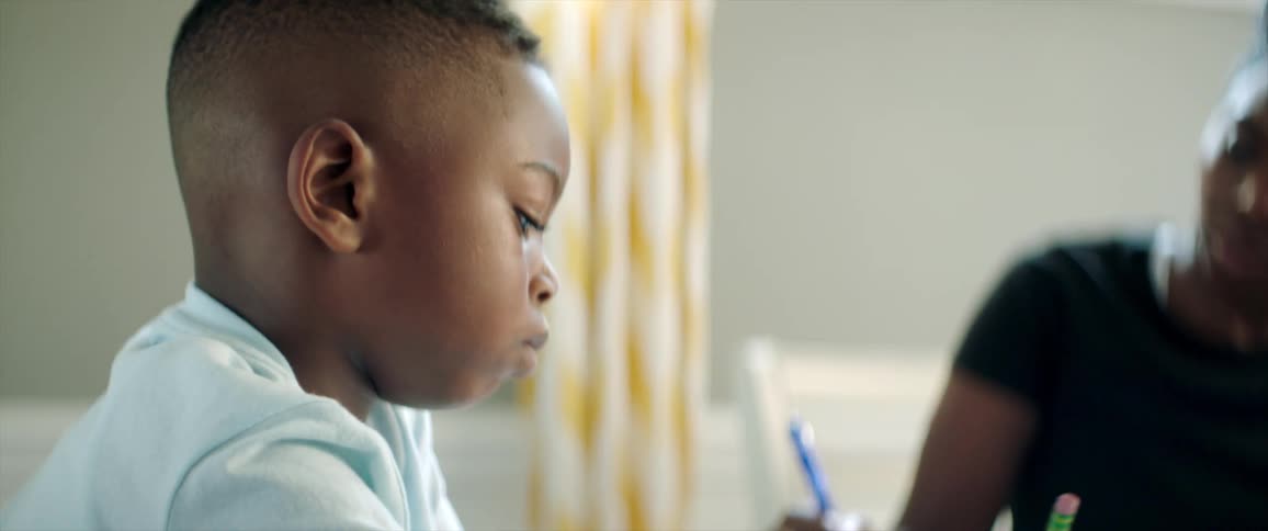 A boy sitting at a table and writing on a book with a pencil next to a woman with a laptop