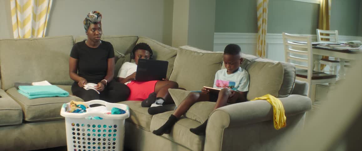 A woman folds laundry while sitting on a sofa next to her sons who are focused on electronics