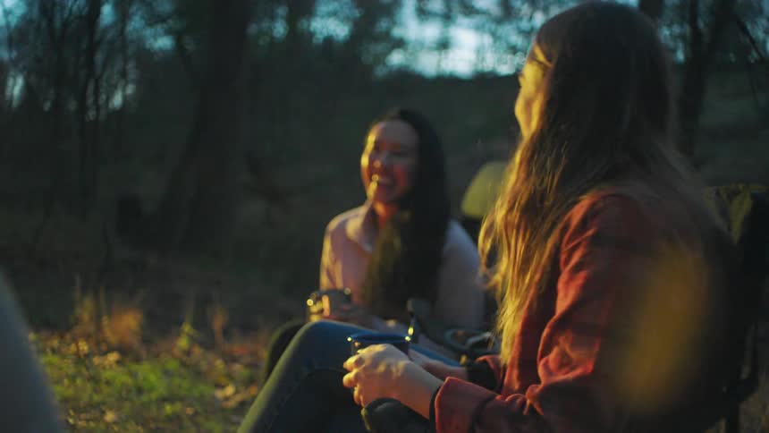 Three women talk and laugh with drinks by the campfire