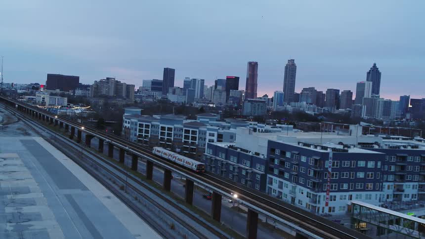Public train on overhead tracks parallels street passing by neighborhood with red sky showing through city skyline