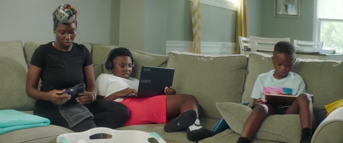 A woman sits on the couch and folds laundry while her boys sit and use a laptop and a tablet
