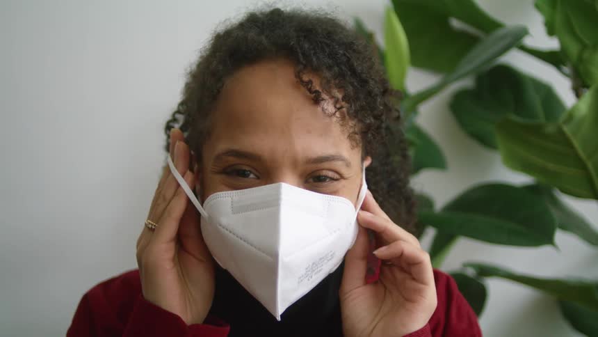 Smiling woman backed by large leafed houseplant removes face mask