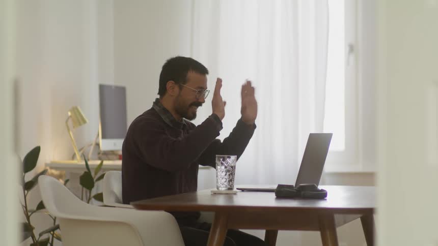 Bearded man sitting in a desk in his home office greeting people on a video call through a laptop
